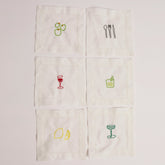 A set of white cocktail napkins with various embroidered designs including a glass of red wine, a lemon, an orange, a cucumber, and a cocktail glass.